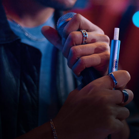 Ein Mann, der in der einen Hand ein Mikrofon und in der anderen ein IQOS hält.