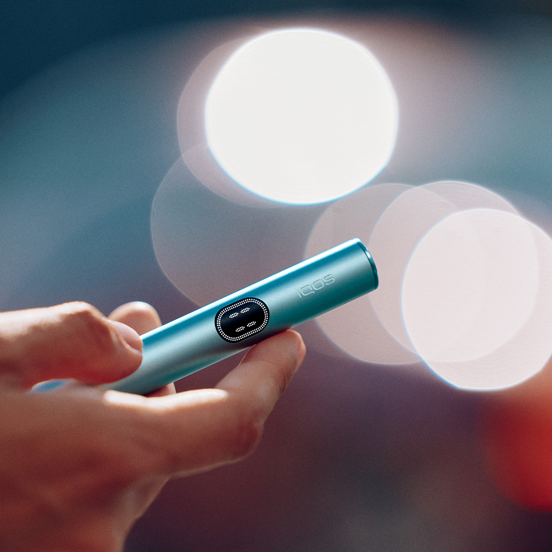 Persona sosteniendo IQOS Iluma i turquesa con luces urbanas desenfocadas