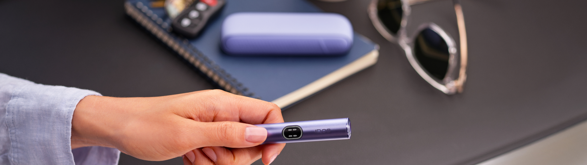 Persona usando IQOS Iluma i turquesa sobre mesa metálica junto a libreta y lentes