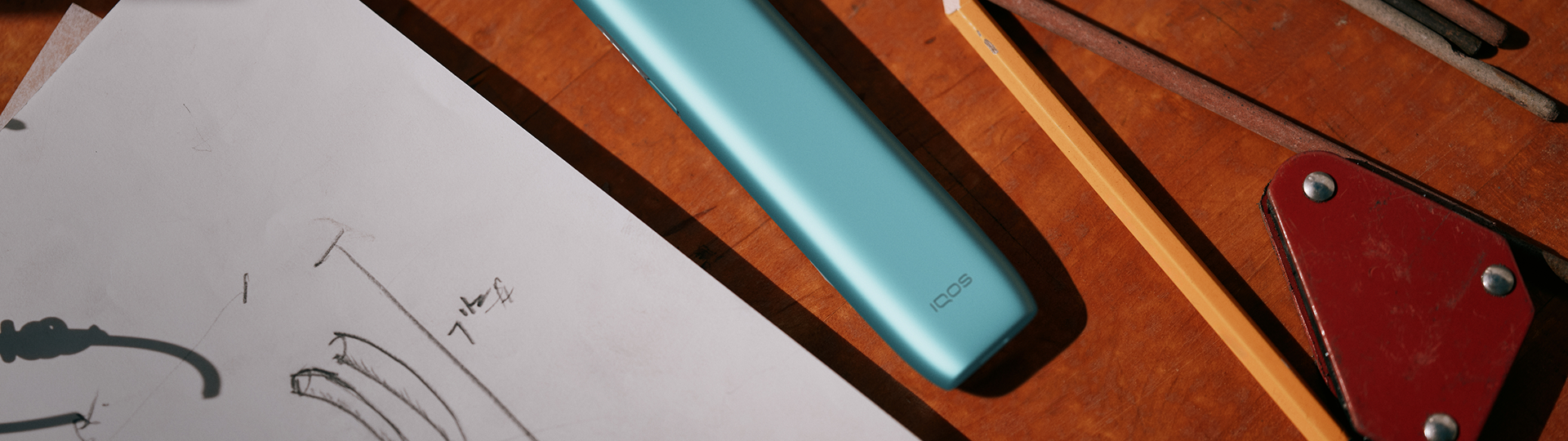 IQOS Iluma i ONE turquesa sobre boceto técnico y mesa de herramientas