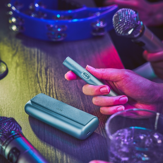 Ruka držící modré zařízení IQOS ILUMA i PRIME; na stole leží nabíječka a mikrofony