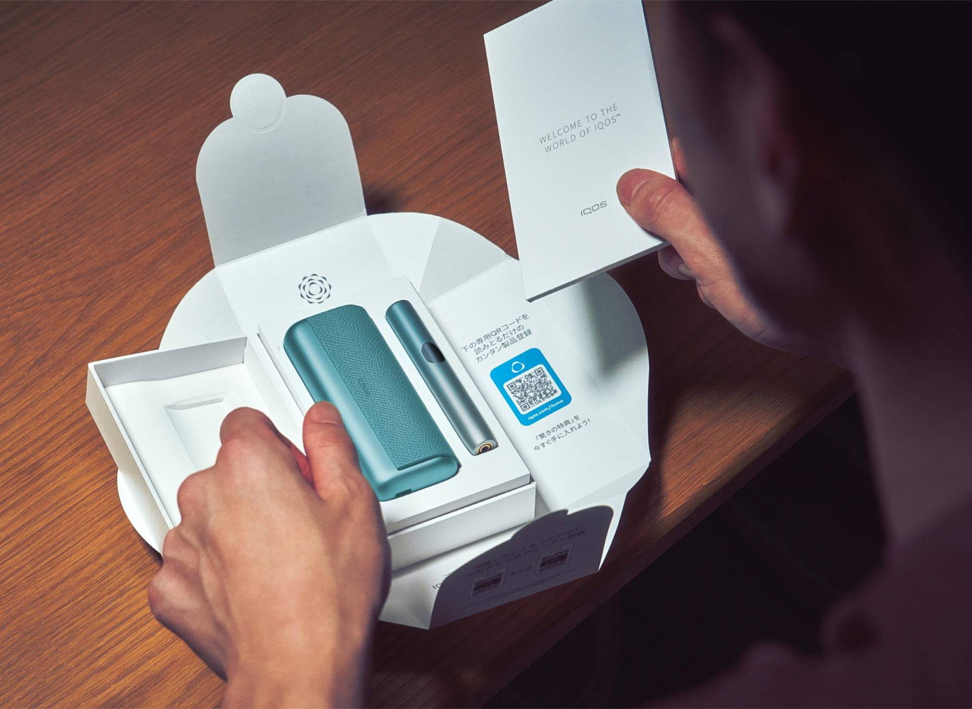 Detail rukou vybalujících modré zařízení IQOS ILUMA i PRIME v pozadí pracovní stůl